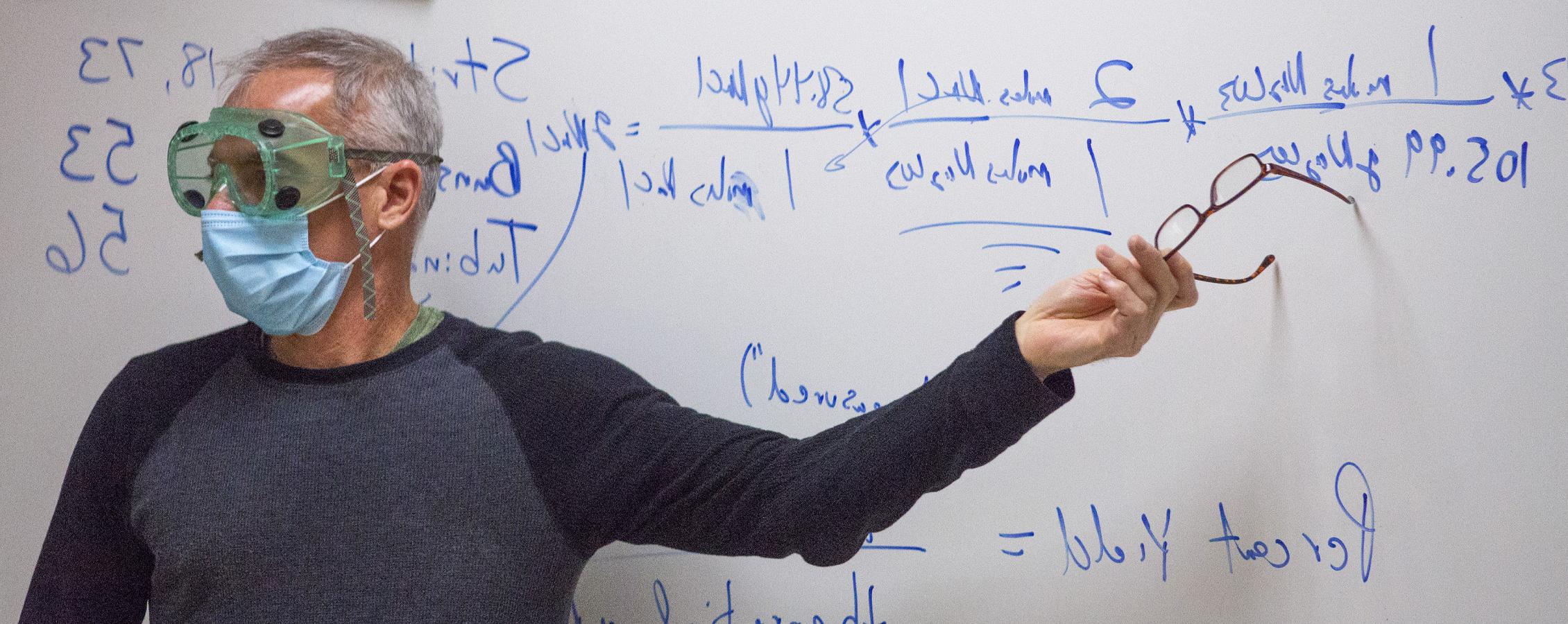 A faculty member, wearing safety googles and a face mask, points at a formula written on a whiteboard.